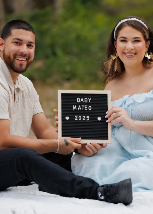 Couple sitting on a blanket, holding a letter board that announces "Baby Mateo 2025" with decorative hearts