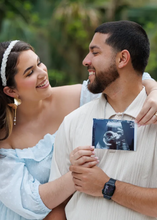 A couple joyfully holds an ultrasound image outdoors, celebrating their upcoming arrival, surrounded by greenery.